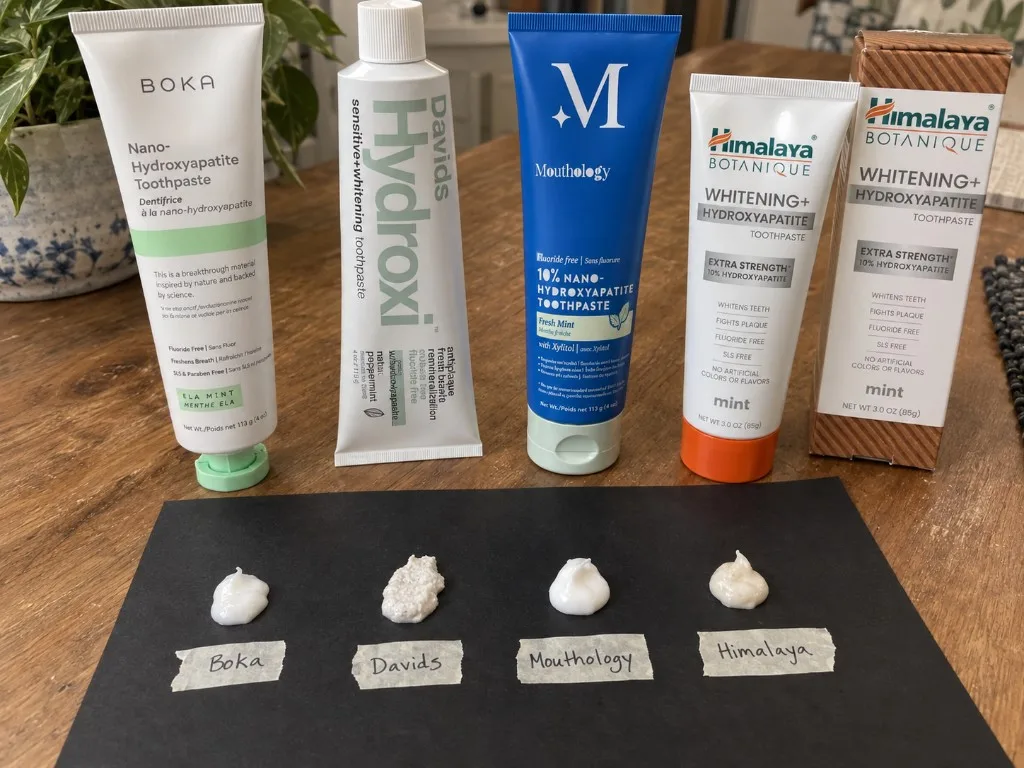Boka Ela Mint, Davids Hydroxi, Mouthology Fresh Mint, and Himalaya Whitening+ Extra Strength tubes on a wood table, with four paste dollops on black paper labeled Boka, Davids, Mouthology, and Himalaya